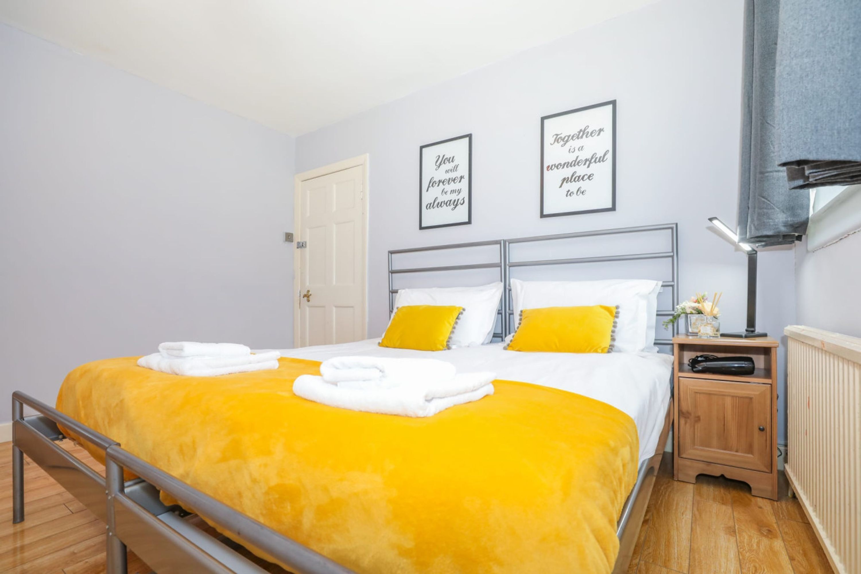 A hotel room with a bed, two bedside tables, a nightstand, a radiator, and two framed quotes on the wall.