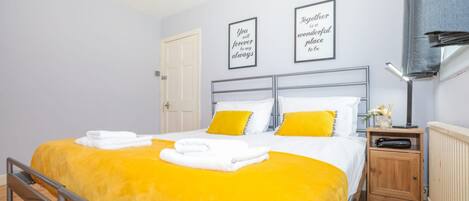 A hotel room with a bed, two bedside tables, a nightstand, a radiator, and two framed quotes on the wall.