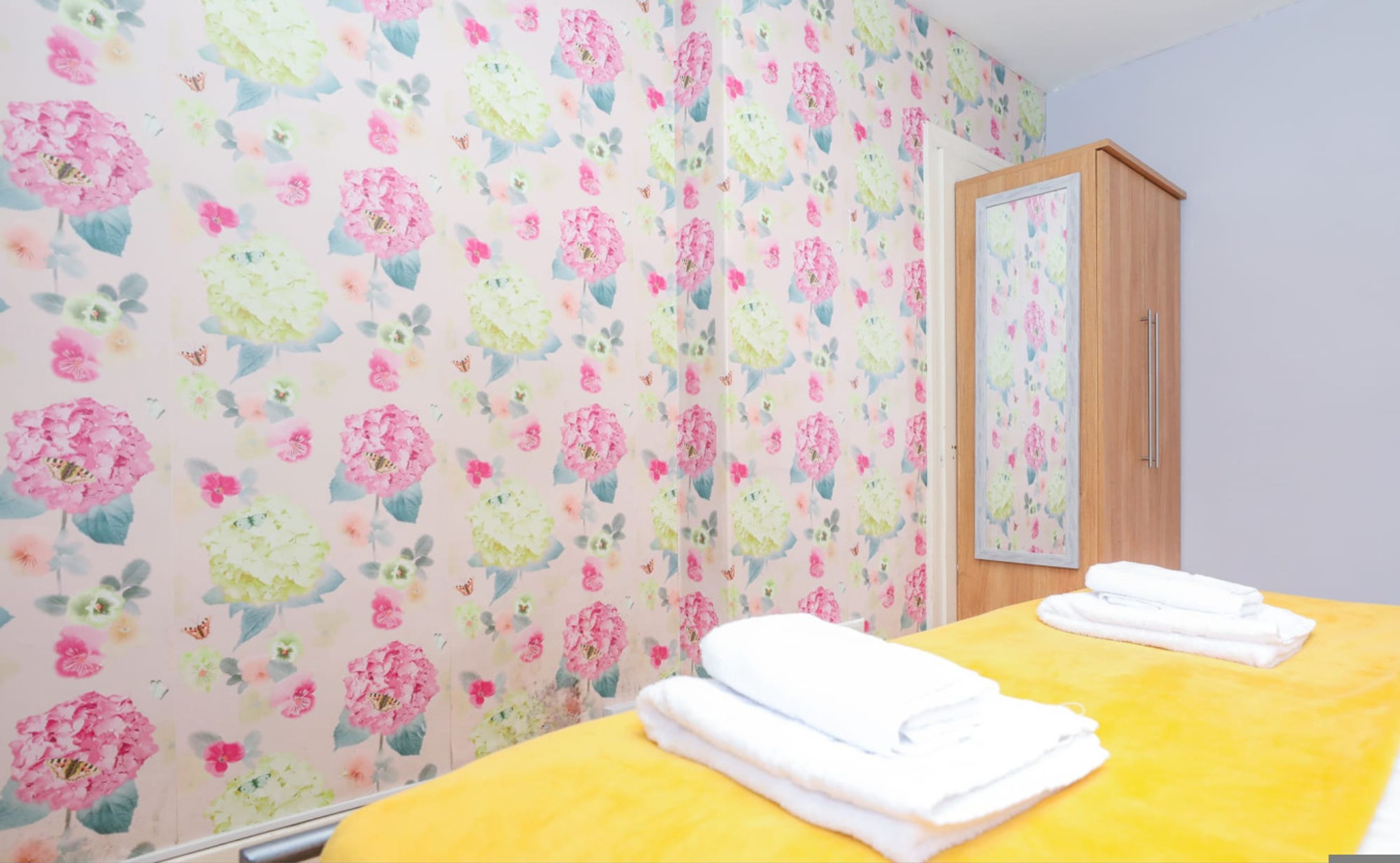 A room with floral wallpaper, a wooden cabinet, and two neatly folded white towels on a yellow bed.