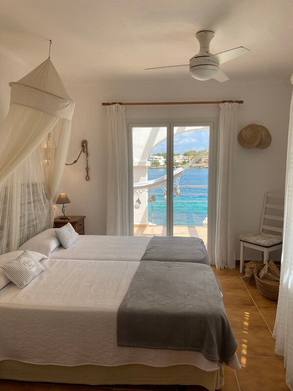 A bedroom with two beds, a ceiling fan, a nightstand, a lamp, a view of the sea, and a chair.