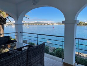 A balcony with wicker furniture, a railing, and a view of a body of water with boats and a distant shoreline.