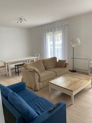 A living room with a sofa, a blue chair, a wooden coffee table, a dining table with chairs, a ceiling fan, and a floor lamp.
