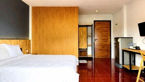 A hotel room with a bed, a ceiling fan, a wooden panel wall, a wooden door, a refrigerator, a TV, and a small table with a chair. - Henry's House (Wichit)