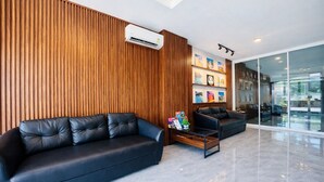 A modern lobby with a wooden panel wall, a black leather sofa, a glass partition, and a mounted air conditioning unit. - Henry's House (Wichit)