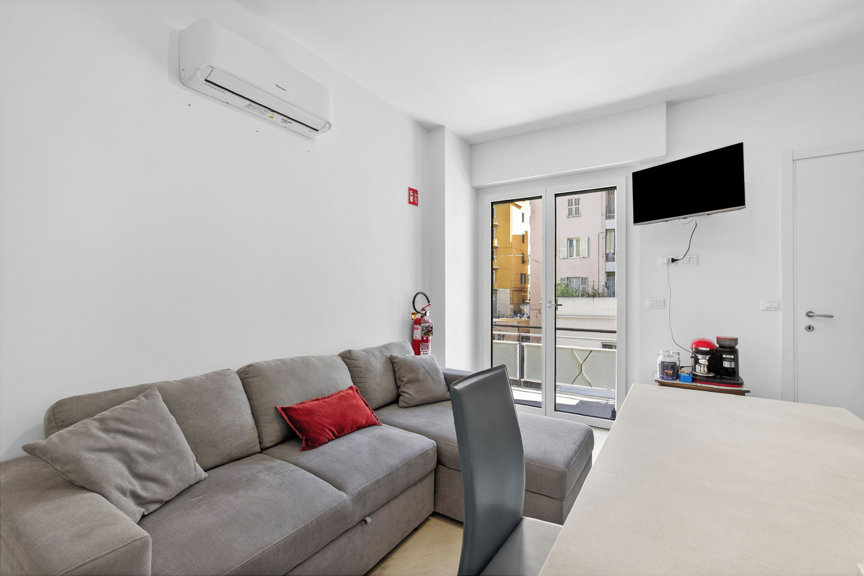 Un salon moderne avec un canapé gris, un coussin rouge, un téléviseur à écran plat et une machine à café posée sur une table.
