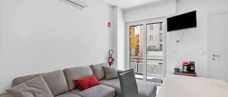 Un salon moderne avec un canapé gris, un coussin rouge, un téléviseur à écran plat et une machine à café posée sur une table.