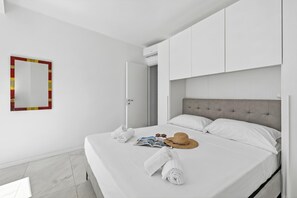 A neatly made bed with towels, a hat, and sunglasses, a mirror with a red frame, and a door leading to another room. - Emilia Sweet - With Balcony + AC (Sanremo)