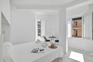 A white room with a bed, a magazine, sunglasses, a hat, and a small table with a chair. - Emilia Sweet - With Balcony + AC (Sanremo)