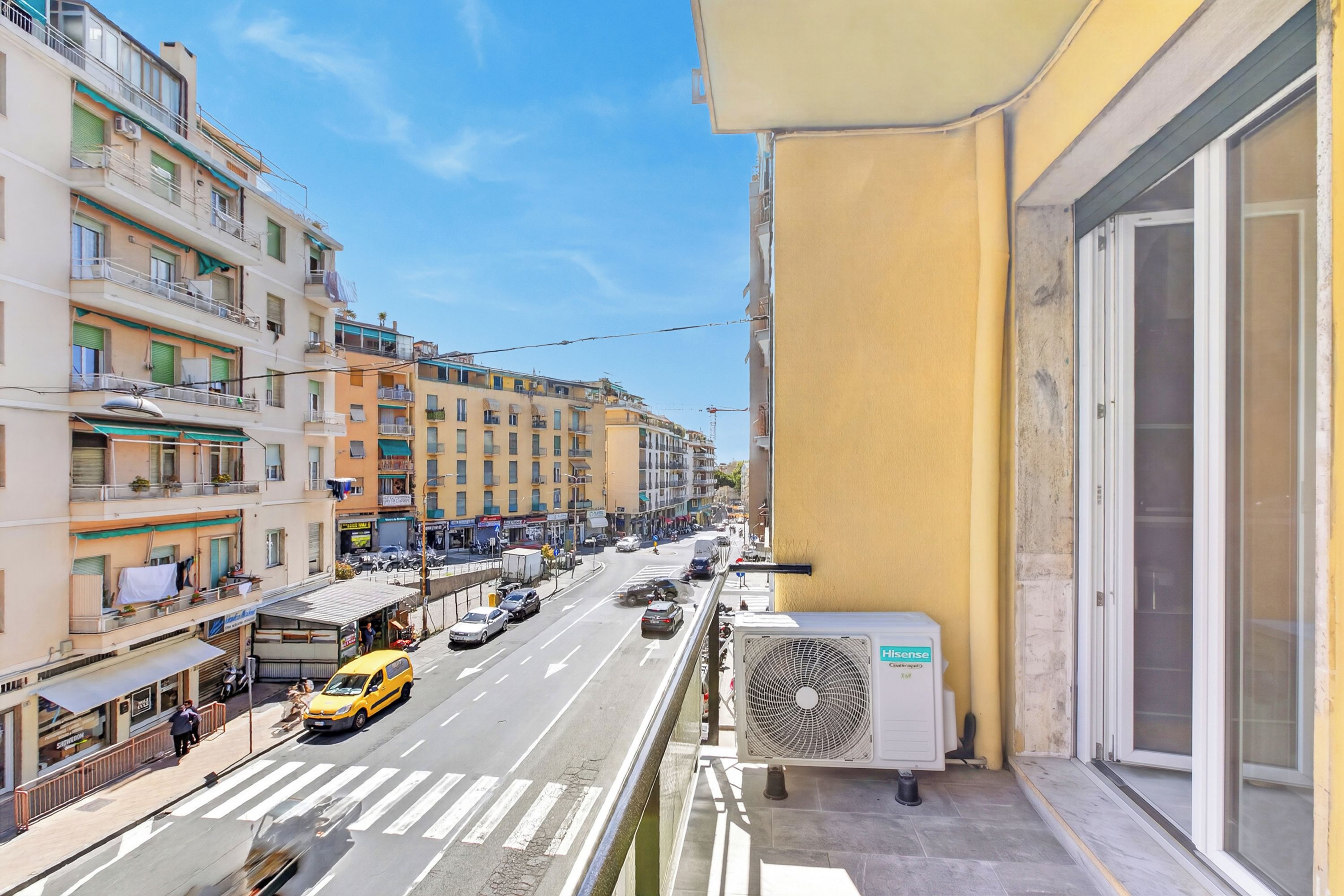 Un balcon donnant sur une rue animée, un immeuble jaune et un appareil de climatisation.