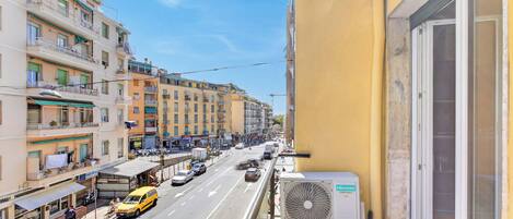 Un balcon donnant sur une rue animée, un immeuble jaune et un appareil de climatisation.