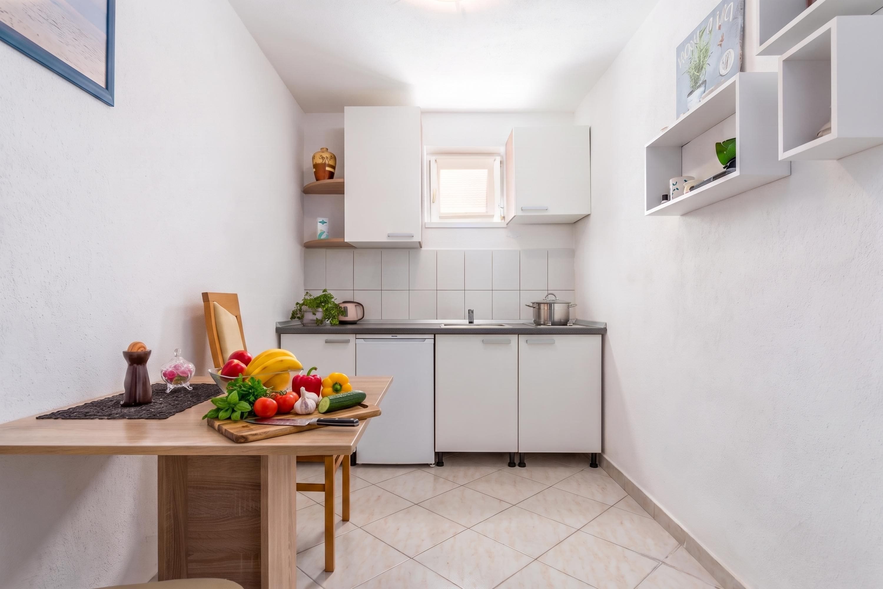 A compact kitchen with white cabinets, a small window, and a dining area with a wooden table set with fruits and vegetables.