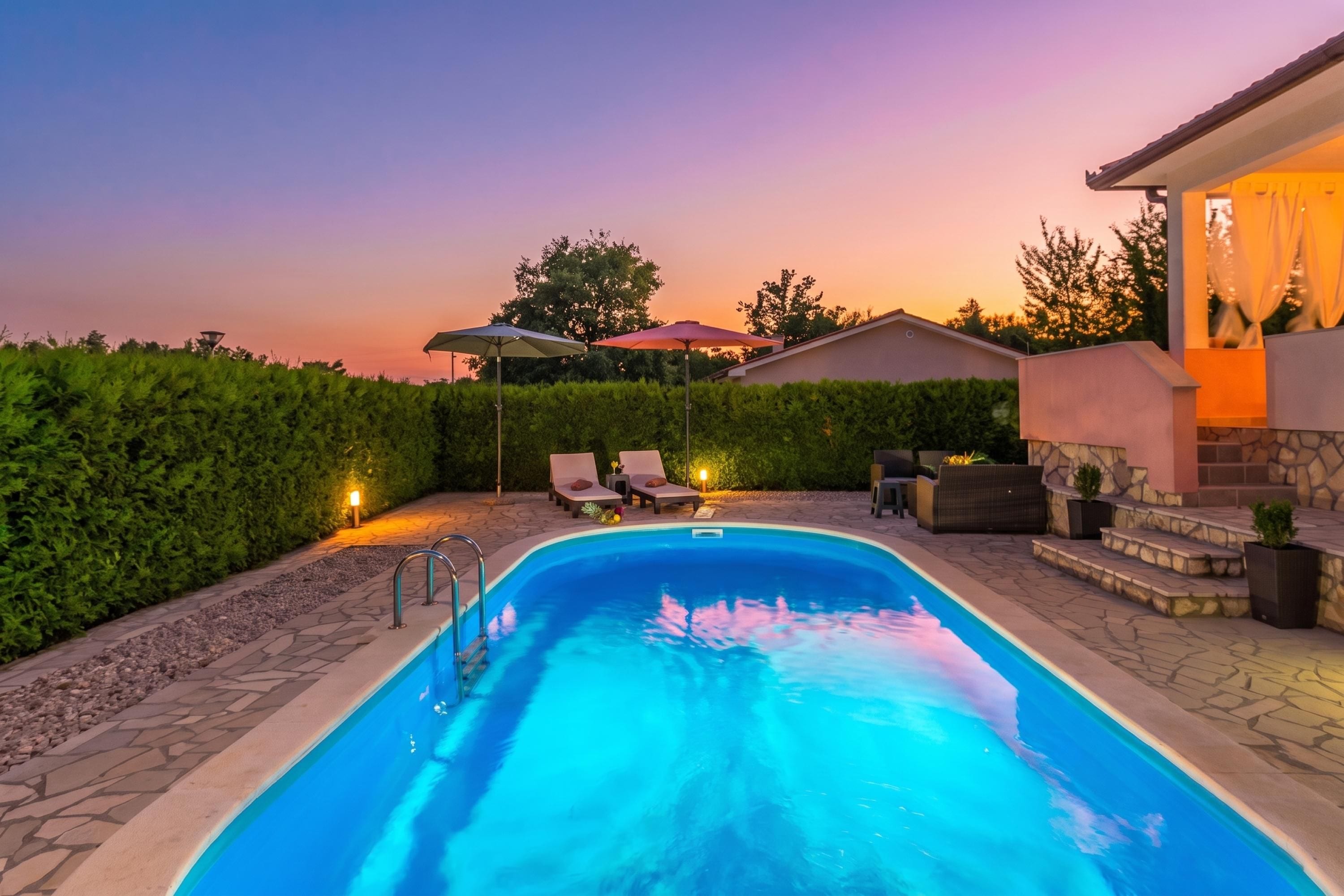 Une piscine entourée d’une terrasse en pierre et d’une haie, avec des chaises longues et des parasols à proximité.