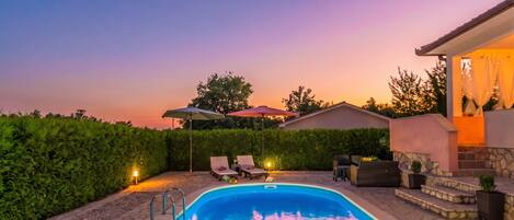 Une piscine entourée d’une terrasse en pierre et d’une haie, avec des chaises longues et des parasols à proximité.