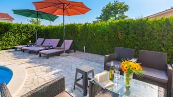A poolside area with lounge chairs, umbrellas, a glass table with a book and wine bottle, and a flower vase.
