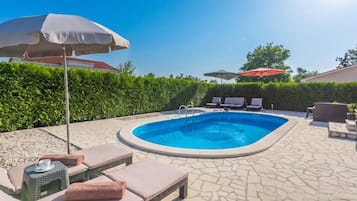 A swimming pool with lounge chairs and umbrellas, surrounded by a hedge and stone flooring.