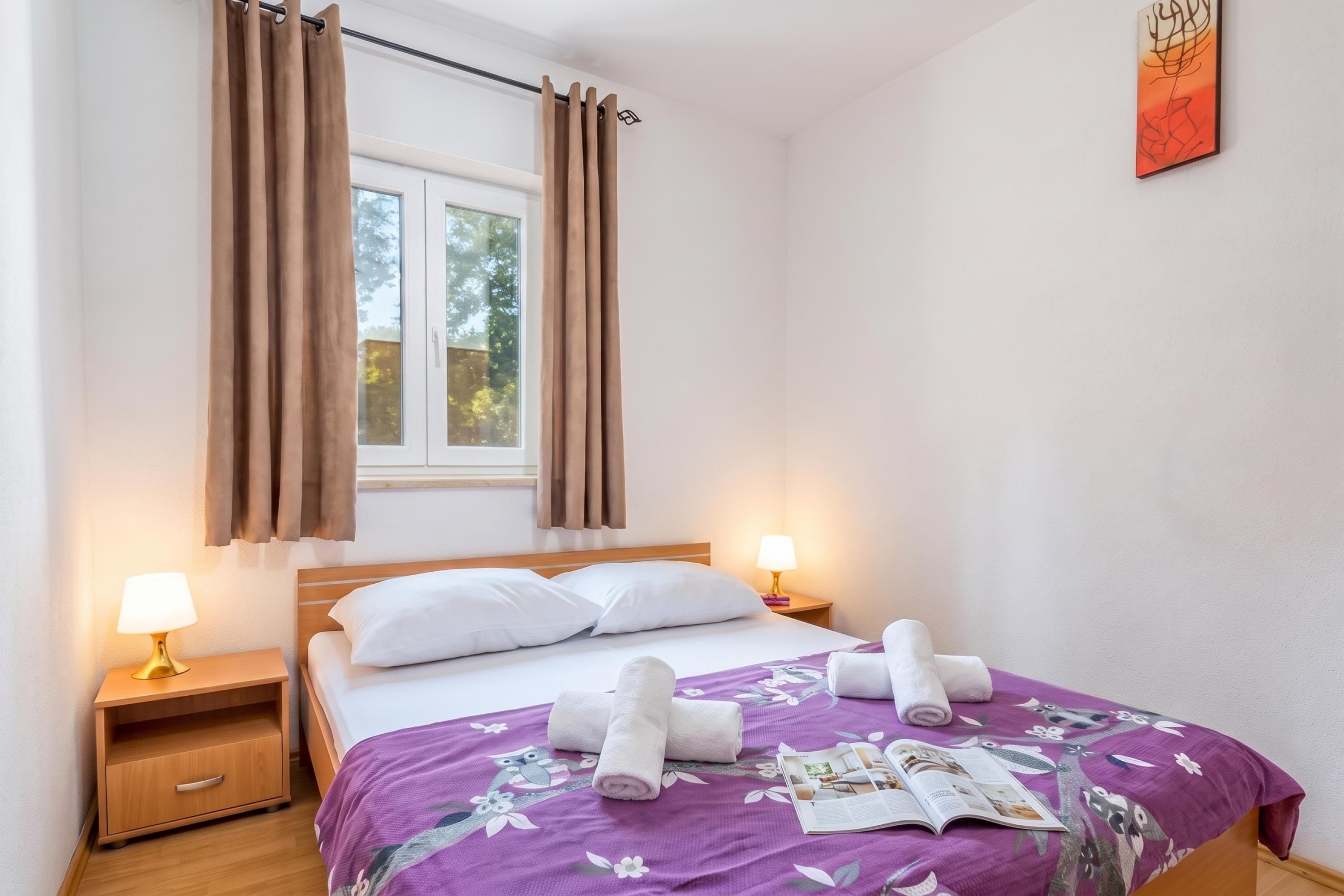 A hotel room with a bed, two bedside lamps, a window with curtains, a wooden nightstand, and a wall art piece.