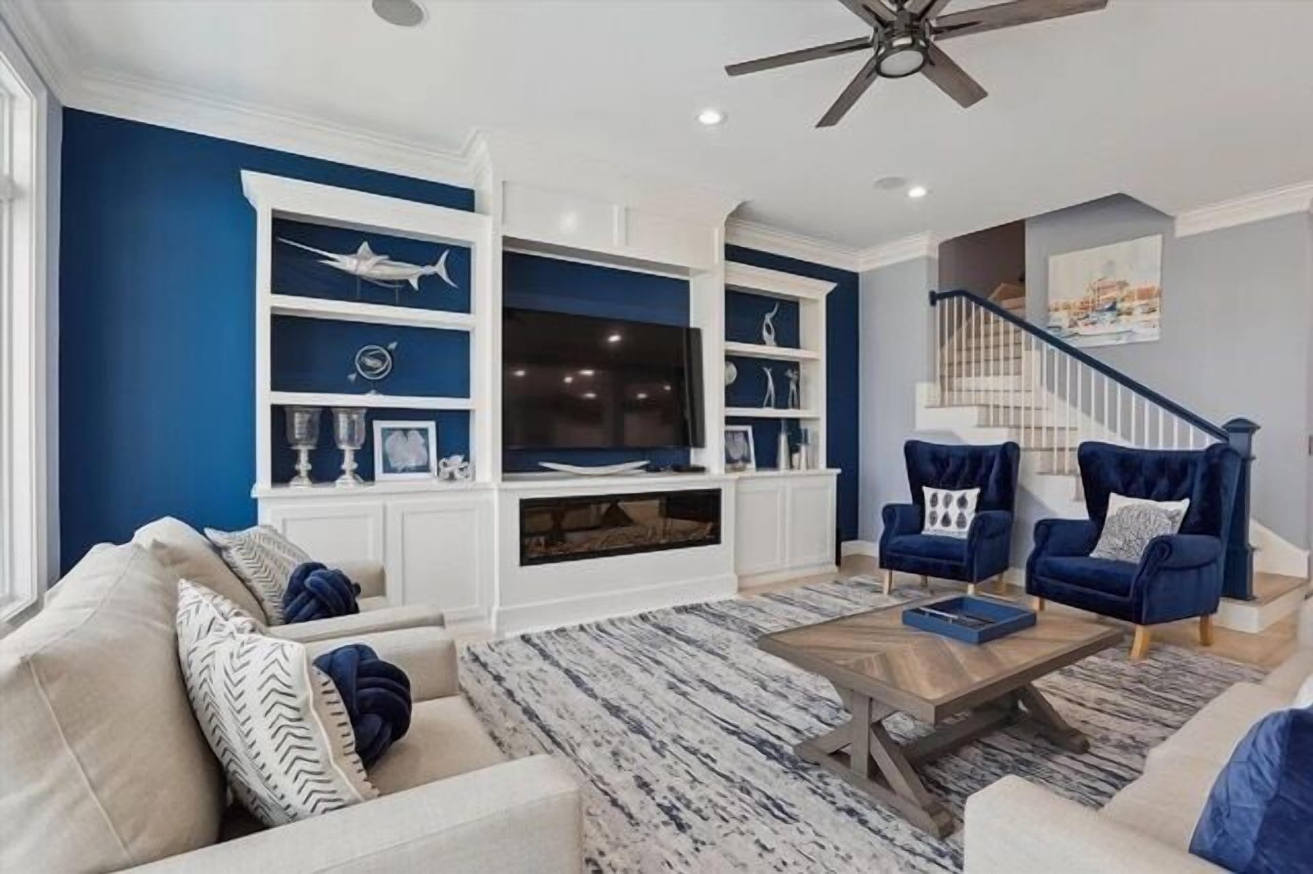 A living room with a blue accent wall, white built-in shelving, a ceiling fan, and a staircase.