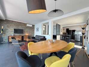 A modern living room with a round wooden table, yellow and blue chairs, a sofa, a TV mounted on a brick wall, and a kitchen in the background. - Agi Joan Margarit 24 Casa for rent in Roses Girona. (Roses)