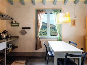 Eine Küche mit Herd, Wasserkocher und Kochgeschirr, ein Esstisch mit Stühlen, ein Fenster mit Vorhängen und eine Hängelampe.
