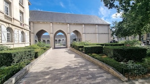 Un chemin pavé mène à un bâtiment doté d’entrées en arc de cercle, entouré de haies et d’arbres bien entretenus.