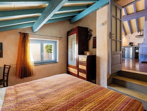 Ein Schlafzimmer mit Bett, Kommode, Fenster mit Vorhängen und Blick auf eine Küche und eine Treppe.