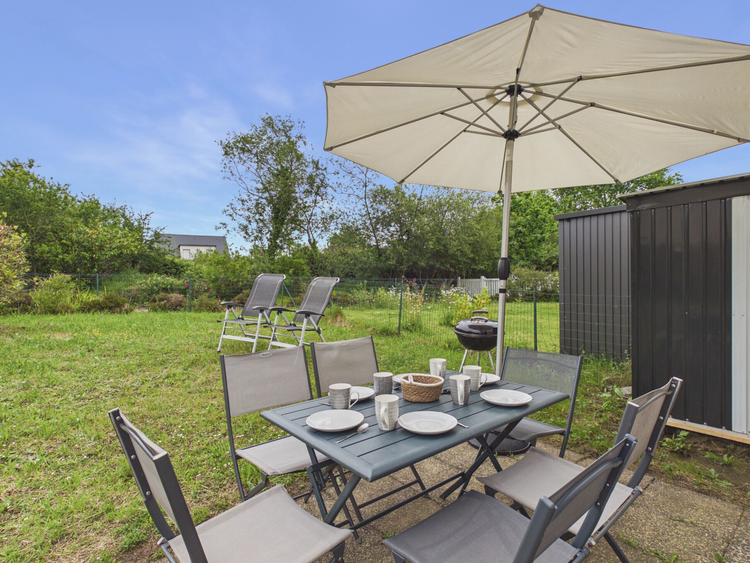 Um pátio externo com uma mesa posta para quatro pessoas, rodeado por cadeiras, sob um grande guarda-sol.