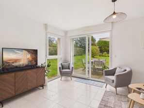 A modern living room with a TV, a grey armchair, a wooden cabinet, and a dining area with outdoor seating. - Near sea – Charming house with garden – Sleeps 6 (Fréhel)