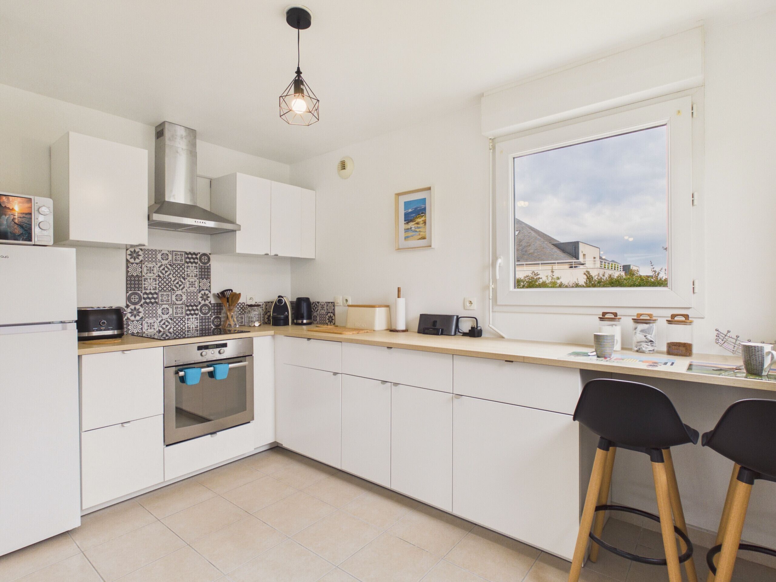 Una cocina moderna con armarios blancos, horno empotrado, microondas, cafetera y ventana con vista a edificios.