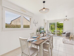 A spacious dining area with a large table, white chairs, and a view through glass doors to an outdoor garden. - Near sea – Charming house with garden – Sleeps 6 (Fréhel)