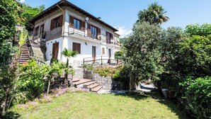 Uma casa de dois andares com varanda, cercada por vegetação exuberante e uma escadaria de pedra que leva à entrada.