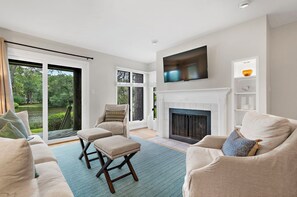 Ein Wohnzimmer mit Kamin, Fernseher und Blick auf einen Garten durch große Fenster.