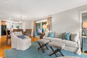 Ein Wohnzimmer mit einer Couch, Sesseln, Poufs, einem Teppich mit Fischgrätmuster und Blick ins Freie durch eine Glastür.