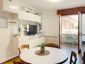 Ein modernes Wohnzimmer mit Fernseher, weißen Möbeln, einem Esstisch mit Stühlen und einem Balkon mit Sitzgelegenheit.