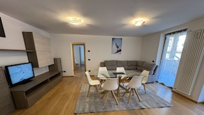 Una sala moderna con televisor, sofá, mesa de comedor con sillas y un ventanal con cortinas.