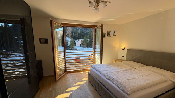 Un dormitorio con cama, mesita de noche, lámpara de pared, ventilador de techo y puerta de cristal corredera que da a un balcón con vistas a un paisaje nevado.
