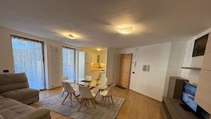 Una sala de estar moderna con un sofá, una mesa de comedor de vidrio con sillas, una zona de cocina y un televisor sobre un mueble.