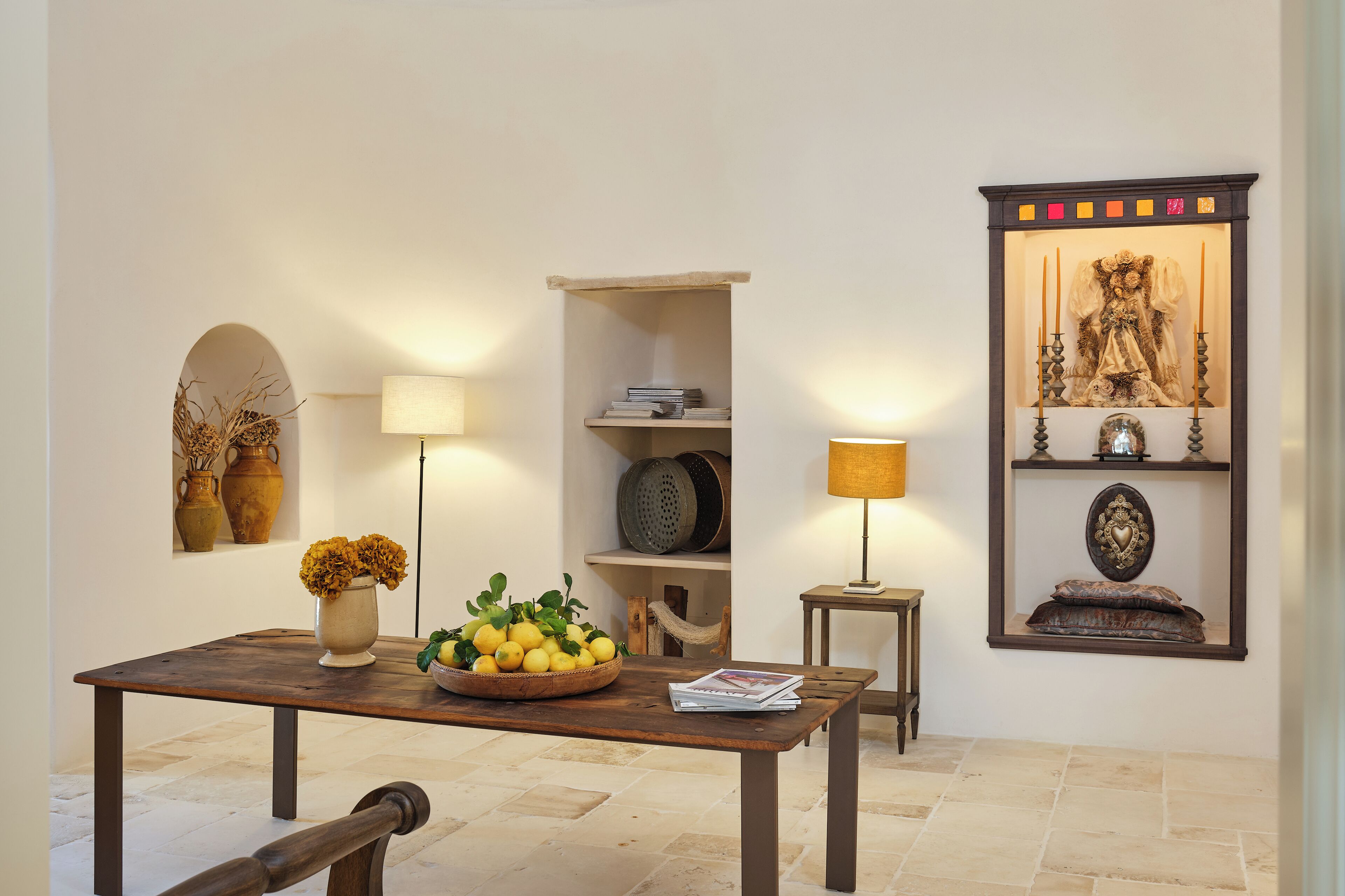 Una habitación con una mesa de madera, un cuenco de limones, un florero con flores, una lámpara, una estantería con libros y un cuadro en la pared.