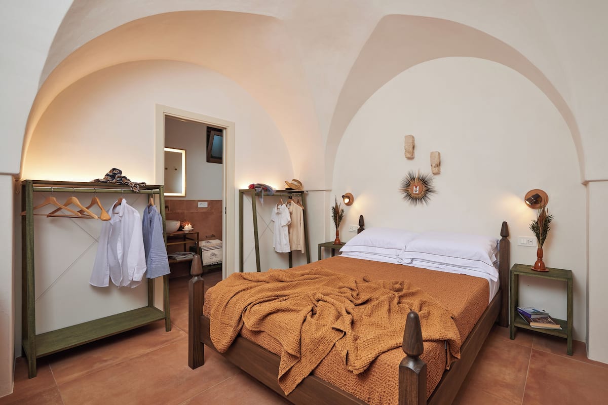 A bedroom with a wooden bed, a hanging clothes rack, a bedside table with a lamp, and a mirror.