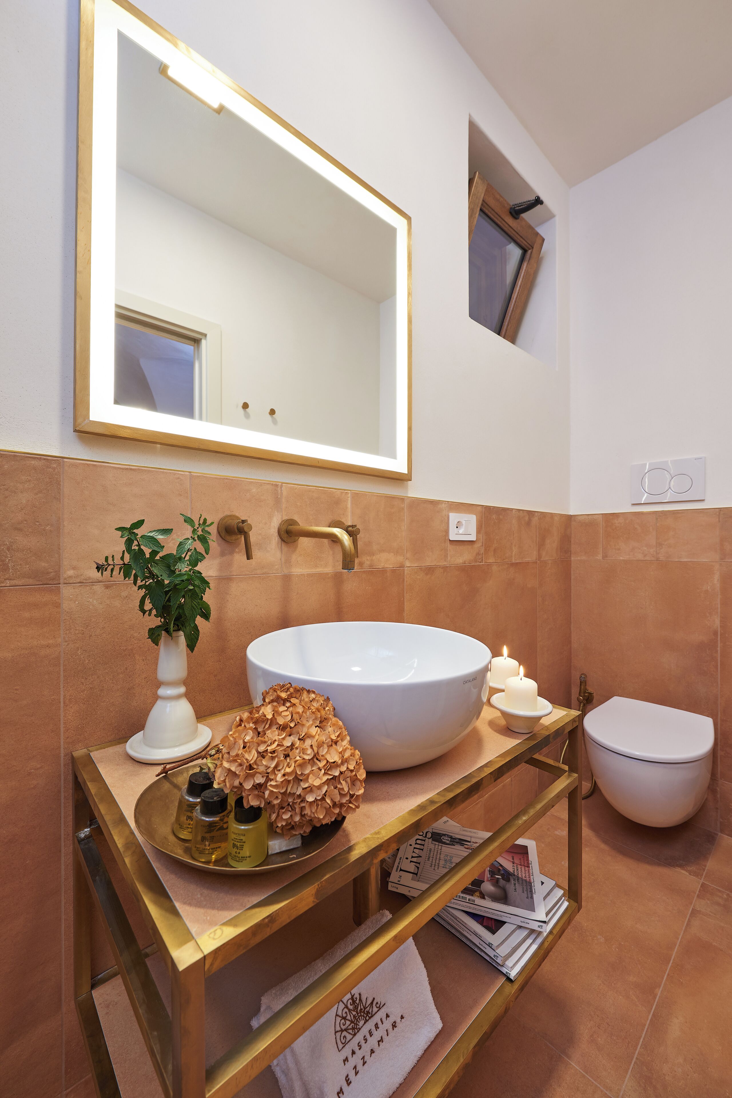 Un baño con un lavabo blanco, un tocador dorado, un espejo, un ventanal pequeño y un inodoro.