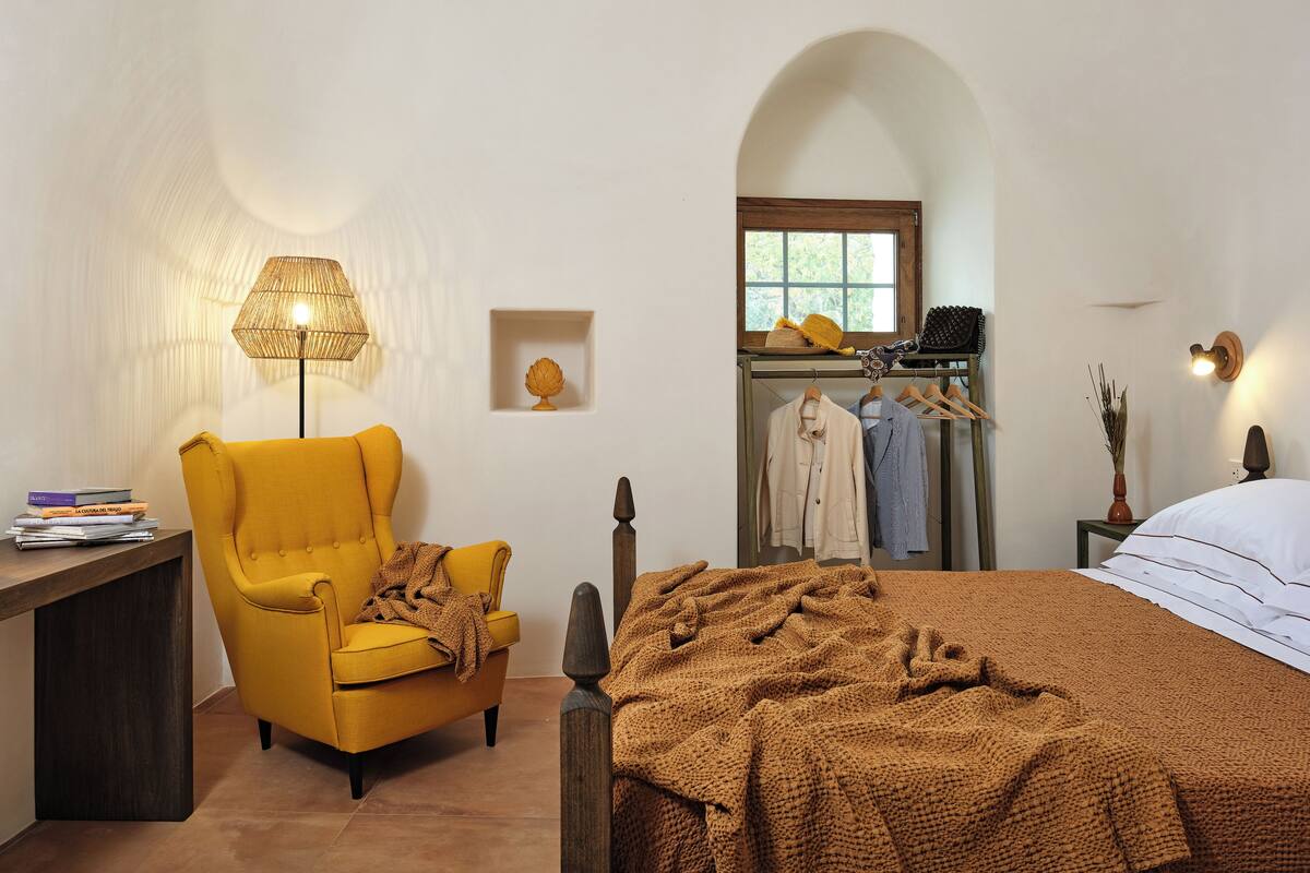 A bedroom with a bed, a chair, a lamp, a shelf with books, and a window.