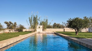Une piscine rectangulaire bordée de pierres, entourée de pelouse et d’arbres.