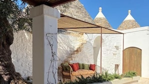 Un espace de restauration en plein air avec une table en pierre, des chaises et un coin salon confortable, offrant une vue sur des bâtiments blancs et un arbre.