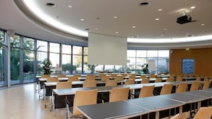 Sala de conferência com mesas, cadeiras, janelas amplas e tela de projeção.