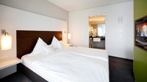 Quarto de hotel com uma cama grande, uma mesinha de cabeceira, um espelho, uma televisão e um banheiro visível por uma porta aberta.