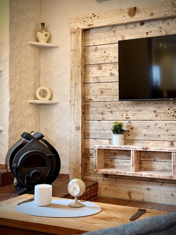 A living room with a wooden wall, a mounted TV, a small shelf with decorative items, and a table with a clock and a mug.