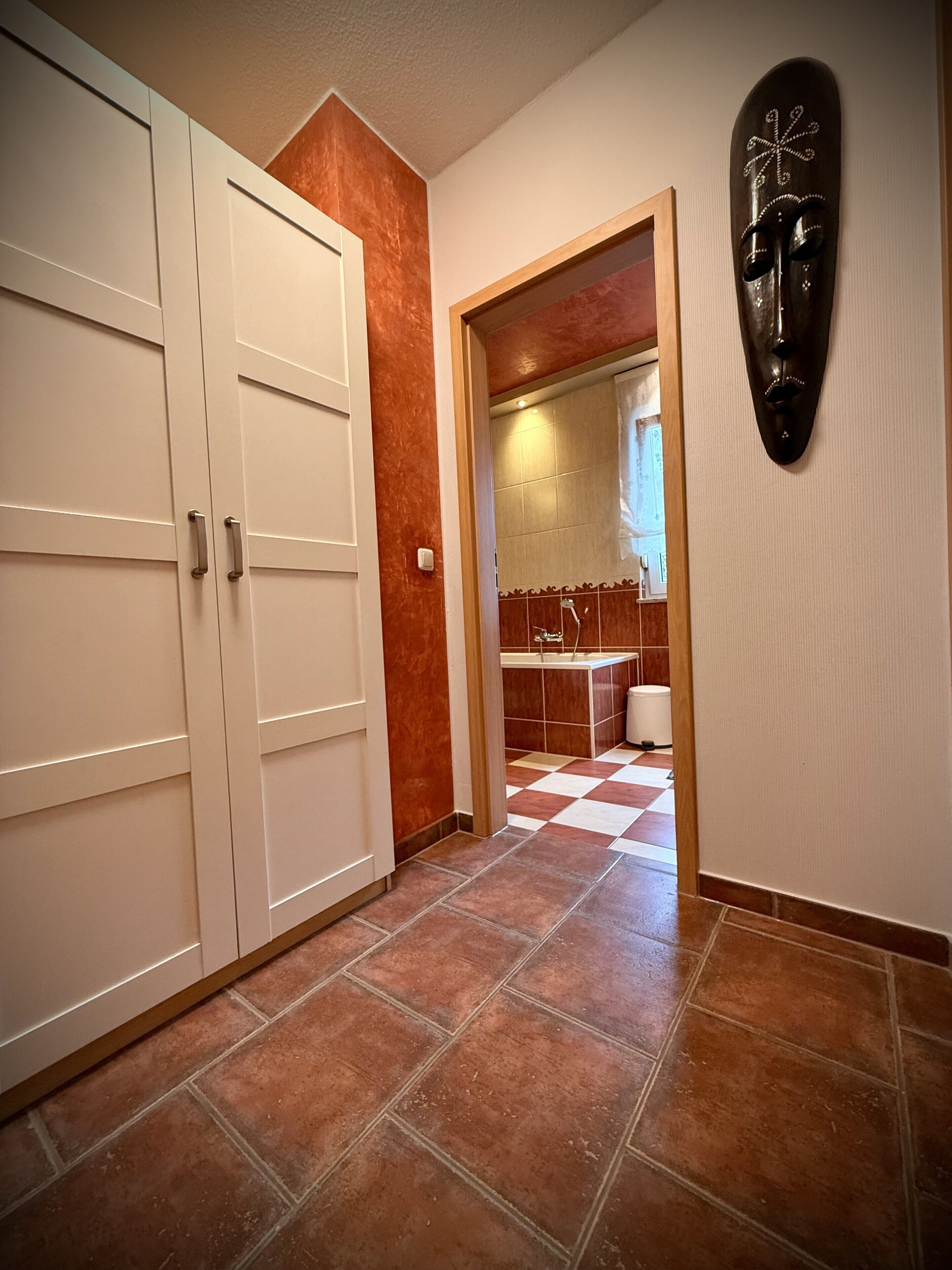 Un corridoio con pavimento a scacchi, una porta bianca e uno specchio che riflette un bagno.