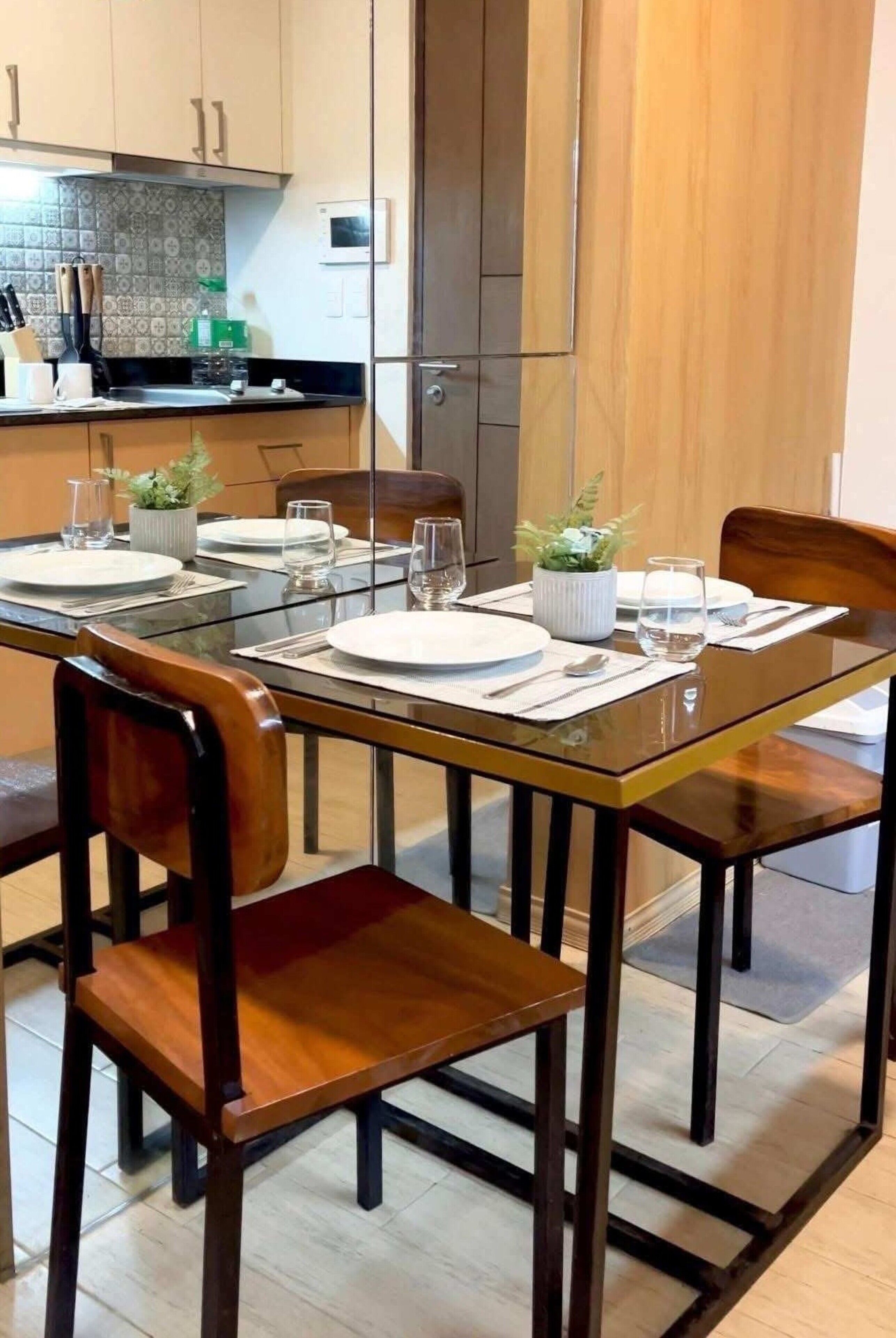 A dining area with a glass table, wooden chairs, and a kitchen in the background.