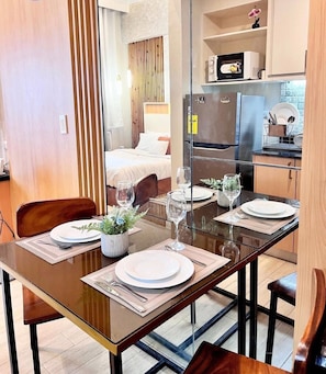 Comedor con una mesa dispuesta para cuatro personas, cocina al fondo y una cama visible a través de una separación.