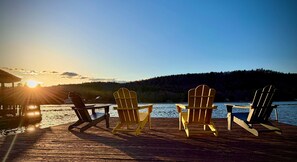 A wooden dock with four Adirondack chairs facing a body of water and a forested hill in the background.