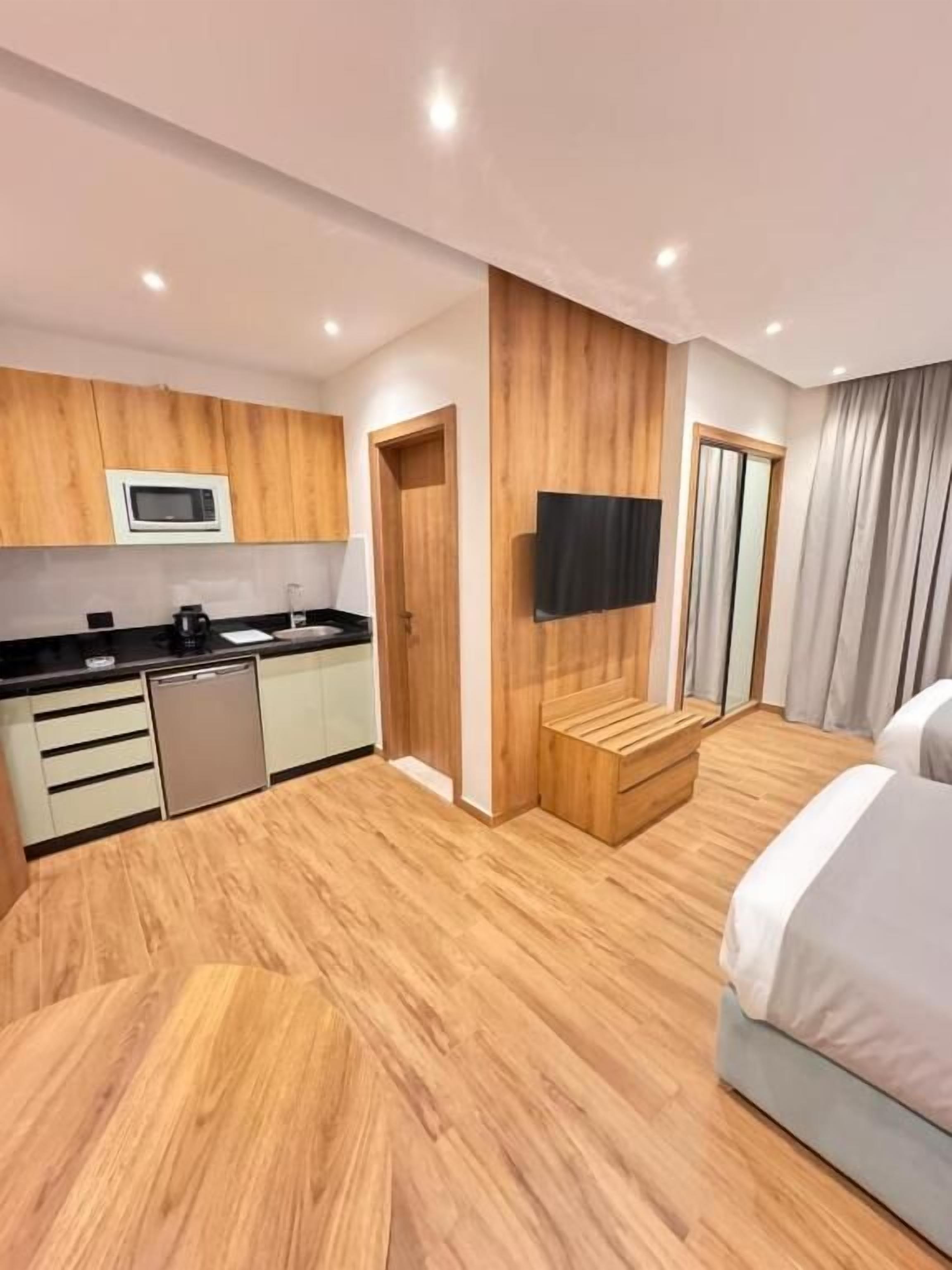 A modern hotel room with a kitchenette, a flat-screen TV, a bed, and a large window.